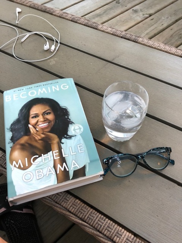 A photo of Michelle Obama's book "Becoming" surrounded by a pair of glasses, a set of wired Apple in-ear headphones, and a glass of water on an outside table.